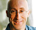 Headshot of a person with glasses, bald head, and a light smile, wearing a blue shirt or jacket.