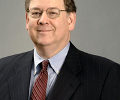 Headshot of a person wearing glasses, a dark suit, blue shirt, and red tie, with a neutral background.