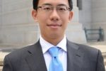 Headshot of a person wearing glasses, a dark suit, white shirt, and blue tie, standing outdoors in front of a stone building.