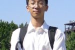 A person wearing glasses and a white shirt, standing outdoors with arms crossed and a backpack, in front of trees and a blue sky.