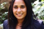 Headshot of a person with long dark hair, wearing a dark blazer and white shirt, smiling outdoors with greenery in the background.