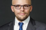 A headshot of a person with glasses and a beard, wearing a suit, white shirt, and blue tie, against a neutral background.