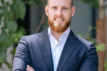 Headshot of a person with a beard, wearing a navy suit and white shirt, standing with arms crossed in an outdoor setting with greenery.