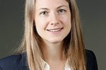 A professional headshot of a person with long hair, wearing a dark blazer over a white shirt, smiling against a neutral background.