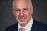 Headshot of a person with short gray hair and a beard, wearing a suit, white shirt, and patterned tie, smiling softly against a dark background.