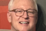 Headshot of a smiling person with light-colored hair and glasses, wearing a light-colored blazer, against a red and dark background.
