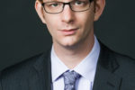 Headshot of a person with short hair, wearing glasses, a dark pinstripe suit, a light-colored shirt, and a paisley tie.