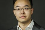 Headshot of a person wearing glasses, a white shirt, and a grey suit jacket, against a dark background.