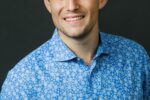 A headshot of a person wearing a blue patterned shirt, smiling against a dark background.