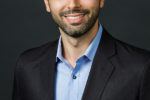 Headshot of a smiling individual with short hair, wearing a dark suit and light blue shirt, set against a dark background.