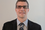 Headshot of a person wearing glasses, a dark suit, a light shirt, and a striped tie, standing against a plain background.