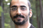 Headshot of a person with short hair, beard, and glasses, wearing a white shirt and dark jacket, with a blurred forest background.