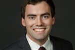 A headshot of a person wearing a dark suit, white shirt, and maroon tie, smiling against a dark background.