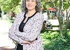 A person with gray curly hair, wearing a patterned blazer and black top, standing outdoors with arms crossed. The setting is a leafy park.