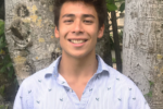 A person smiling outdoors, standing in front of tree trunks, wearing a light blue, patterned shirt.