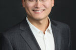 A headshot of a person wearing glasses, a dark suit, and a white shirt, smiling against a black background.