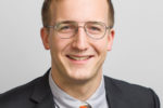 A headshot of a person wearing glasses, a dark suit, a light shirt, and a patterned orange tie, smiling against a plain background.