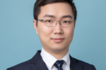 A headshot of a person with short dark hair and glasses, wearing a dark suit, white shirt, and a patterned tie, set against a light blue background.