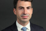 A professional headshot of a person wearing a suit, white shirt, and blue patterned tie against a dark background.