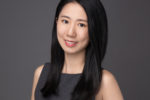 A professional headshot of a person with long black hair, wearing a sleeveless dark dress and smiling against a gray background.