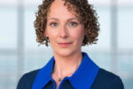 Headshot of a person with curly hair, wearing a blue jacket and a necklace. The background is a blurred grid-like pattern.