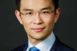 A professional headshot of a person wearing glasses, dressed in a blue suit, white shirt, and blue tie.