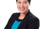 Headshot of a person with short dark hair, wearing a black blazer over a teal top, smiling with arms crossed against a white background.