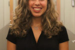 A person with curly hair smiling in an office hallway, wearing a black top.