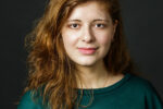 A headshot of a person with long, wavy brown hair, wearing a green top, against a dark background.