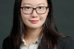 A professional headshot of a person with long dark hair, wearing glasses, a dark blazer, and a white shirt, set against a plain background.