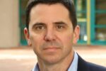 Headshot of a person with short dark hair, wearing a blue collared shirt and dark blazer, standing outside with a blurred background.