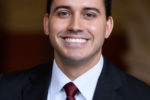 A person in a formal business suit and tie, smiling, against a neutral background. Headshot format.