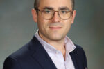 Headshot of a person wearing glasses, a dark blazer, and a light checkered shirt against a plain background.