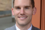 A headshot of a smiling person in a gray suit, white shirt, and striped tie.