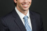 Headshot of a person wearing a dark pinstripe suit, white shirt, and light blue striped tie, smiling against a dark background.