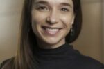 Headshot of a person with long brown hair, smiling, wearing a black turtleneck and earrings.
