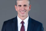 Headshot of person in a suit and tie against a gray background.