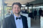 A person in a tuxedo with a bow tie, standing in an indoor setting with a blurred background of people and tables.