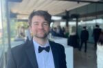 A person in a tuxedo with a bow tie, standing in an indoor setting with a blurred background of people and tables.