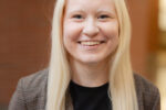 A smiling person with long, platinum blond hair, wearing a brown plaid blazer and a black shirt.