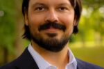 A headshot of a person with shoulder-length dark hair and a beard, wearing a white shirt and dark blazer, standing outdoors with blurred greenery in the background.