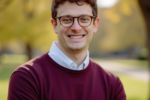 Headshot of a person outdoors, wearing glasses, a maroon sweater, and a collared shirt, smiling with trees in the background.