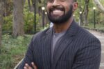 A person with short hair and a beard is smiling, standing outdoors on a walkway lined with trees and lamps. They are wearing a striped suit jacket and have their arms crossed.