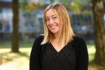 A headshot of a person with shoulder-length blond hair, wearing a black top, smiling outdoors with blurred trees in the background.