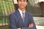 A person in a suit and tie standing with arms crossed outside in front of a glass and brick building.