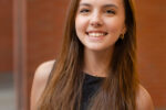 A person with long brown hair and a black top smiling, posing for a headshot against a blurred brown background.