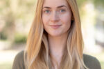 A headshot of a person with long, straight blonde hair, wearing a green top, standing outside with a blurred natural background.