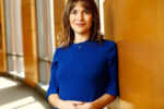 A headshot of a person with shoulder-length hair wearing a blue dress, standing indoors against a wooden wall. The setting is well-lit with natural light.