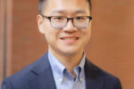 A person wearing glasses, a blue dress shirt, and a dark blazer, smiling in a headshot with a blurred brick background.