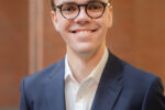 Headshot of a person wearing glasses and a navy suit with a white shirt, smiling in an indoor setting with a brick wall background.